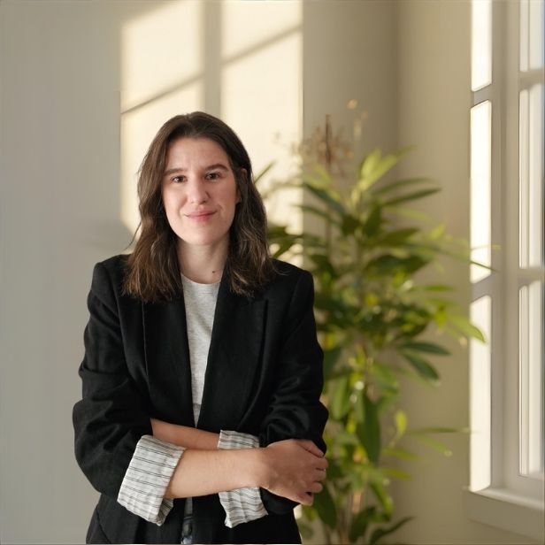 A mature Black male therapist with a salt-and-pepper beard, wearing a green sweater and khakis, sits in a cozy office with a large window and leafy plants. He radiates calm and wisdom.