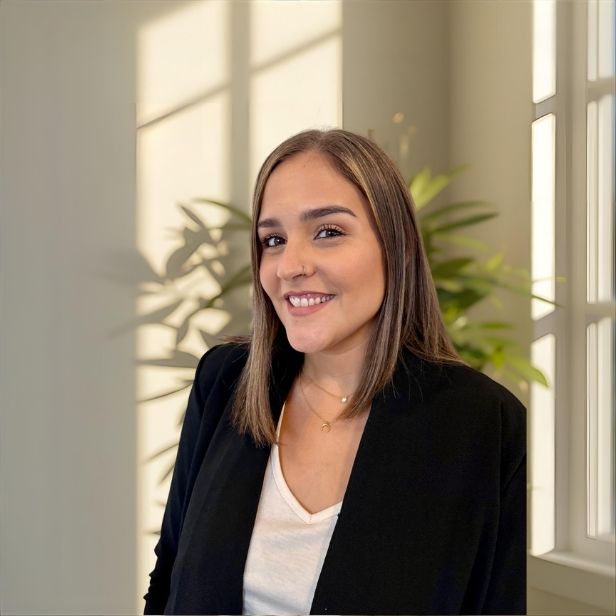 A middle-aged female therapist with short curly hair, wearing glasses and a soft blue blouse, sits in a modern office with bookshelves and plants. She smiles warmly, exuding trust and professionalism.