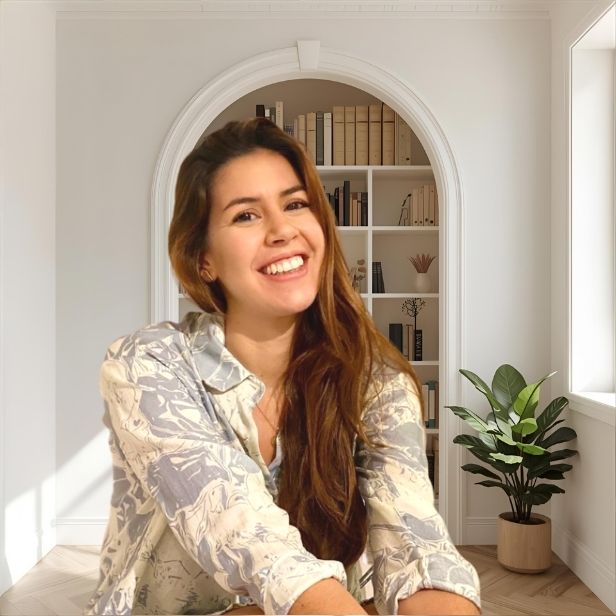 A young male therapist of South Asian descent, wearing a tailored grey suit and open-collar shirt, sits in a bright office with abstract art and a potted plant. He appears attentive and approachable.