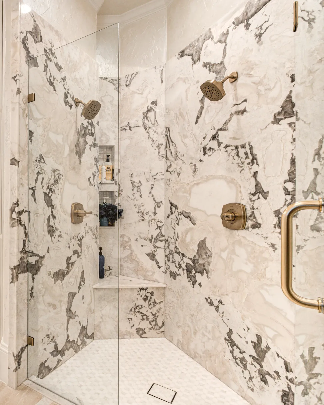 A stunning, spacious walk-in shower adorned with unique white and gray tiles on the walls, featuring two bronze shower heads and a glass divider. The floor is tiled with small white hexagons, complementing the luxurious aesthetic. Two built-in niches provide convenient storage for bath products.