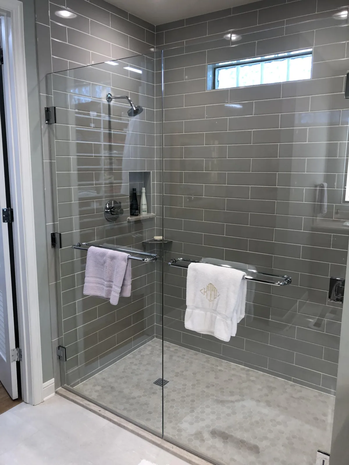 A sleek, modern shower with light gray subway tiles, a glass door, and a matching glass side panel. The shower features a small high window, brushed metal fixtures, and a floor of gray pebble tiles. 