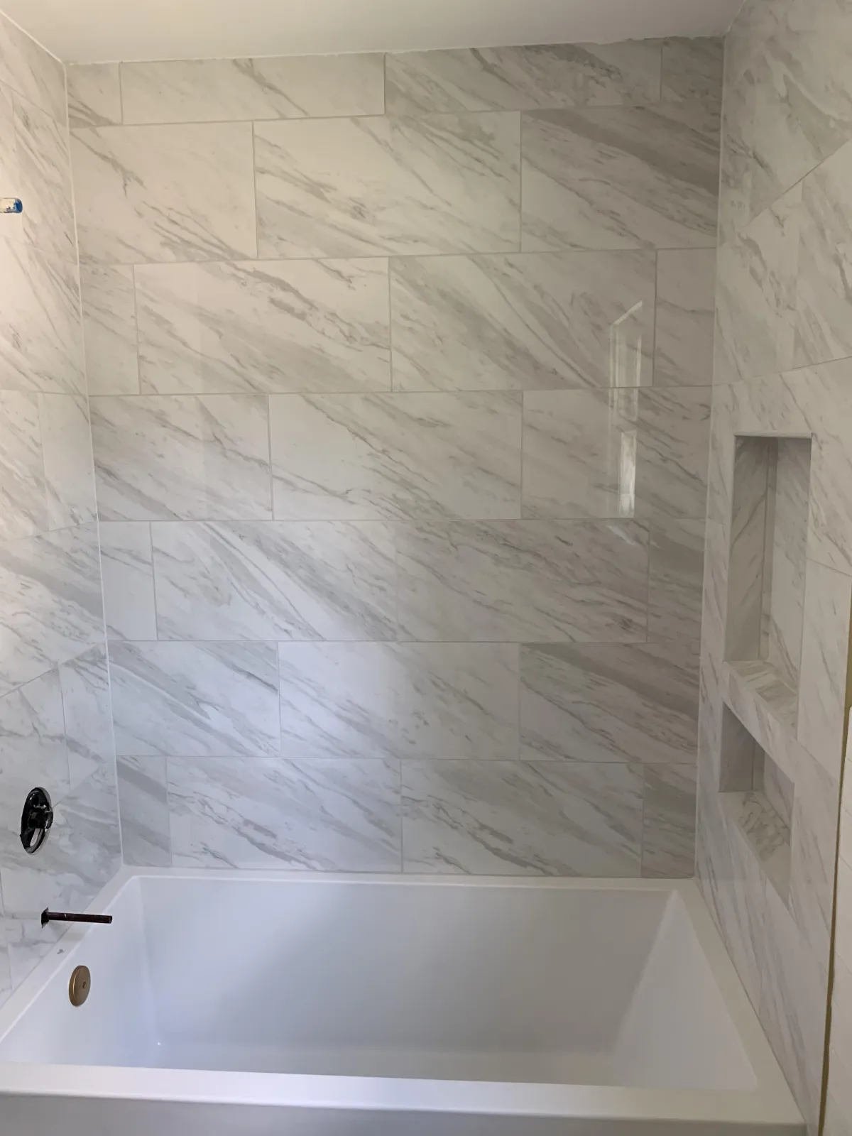A luxurious bathroom with a large built-in bathtub surrounded by walls and a ledge clad in polished marble tiles showcasing varying shades of gray veining. The design includes a wall niche for bath products and a minimalist black faucet.