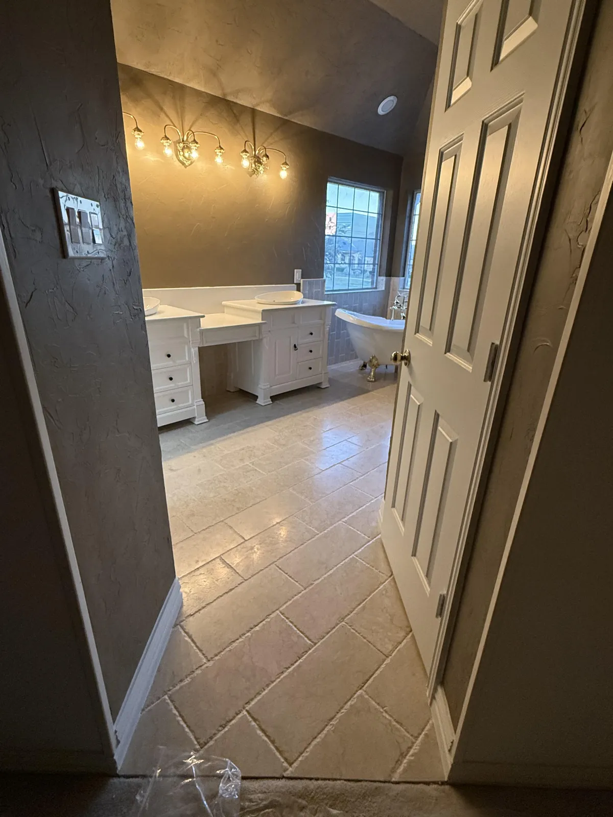 A luxurious bathroom with a large built-in bathtub surrounded by walls and a ledge clad in polished marble tiles showcasing varying shades of gray veining. The design includes a wall niche for bath products and a minimalist black faucet.