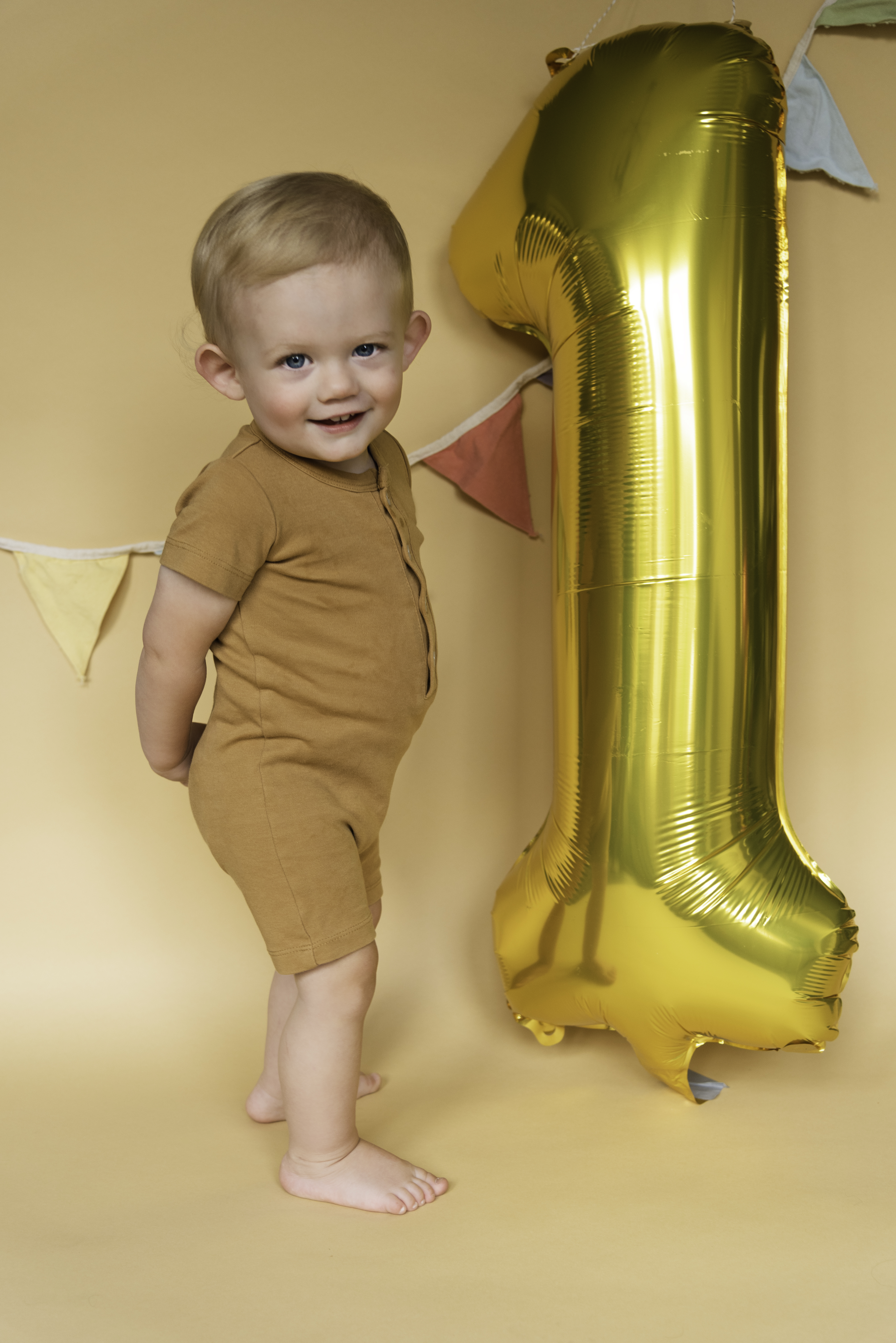 1 year old boy in onesie, smiling with number 1 balloon and banner