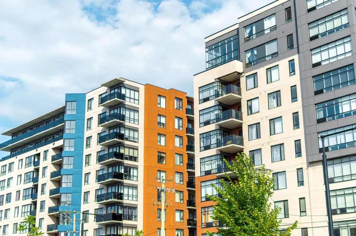 Modern multifamily apartment buildings with mixed brick and glass exteriors under a blue sky, featuring large windows, private balconies, and urban greenery in the foreground.