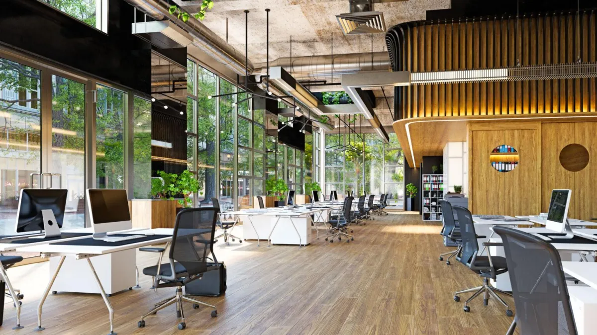 Modern open-concept office with wood flooring, rows of sleek white desks, ergonomic chairs, and desktop computers, surrounded by large floor-to-ceiling windows with natural light and greenery visible outside.