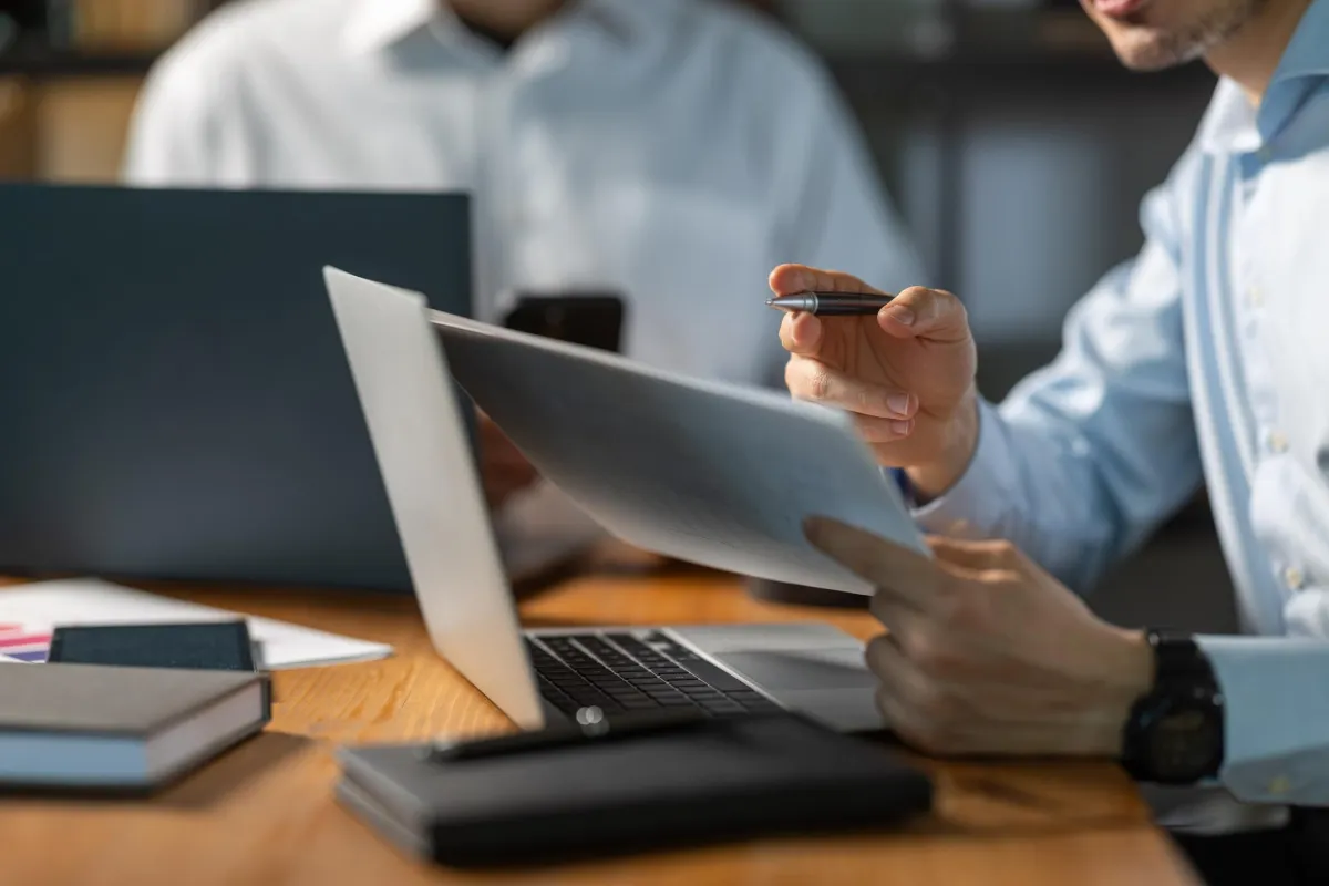Close-up of professionals working together at a desk with a laptop, documents, notebooks, and a pen, discussing business or project details in an office setting.