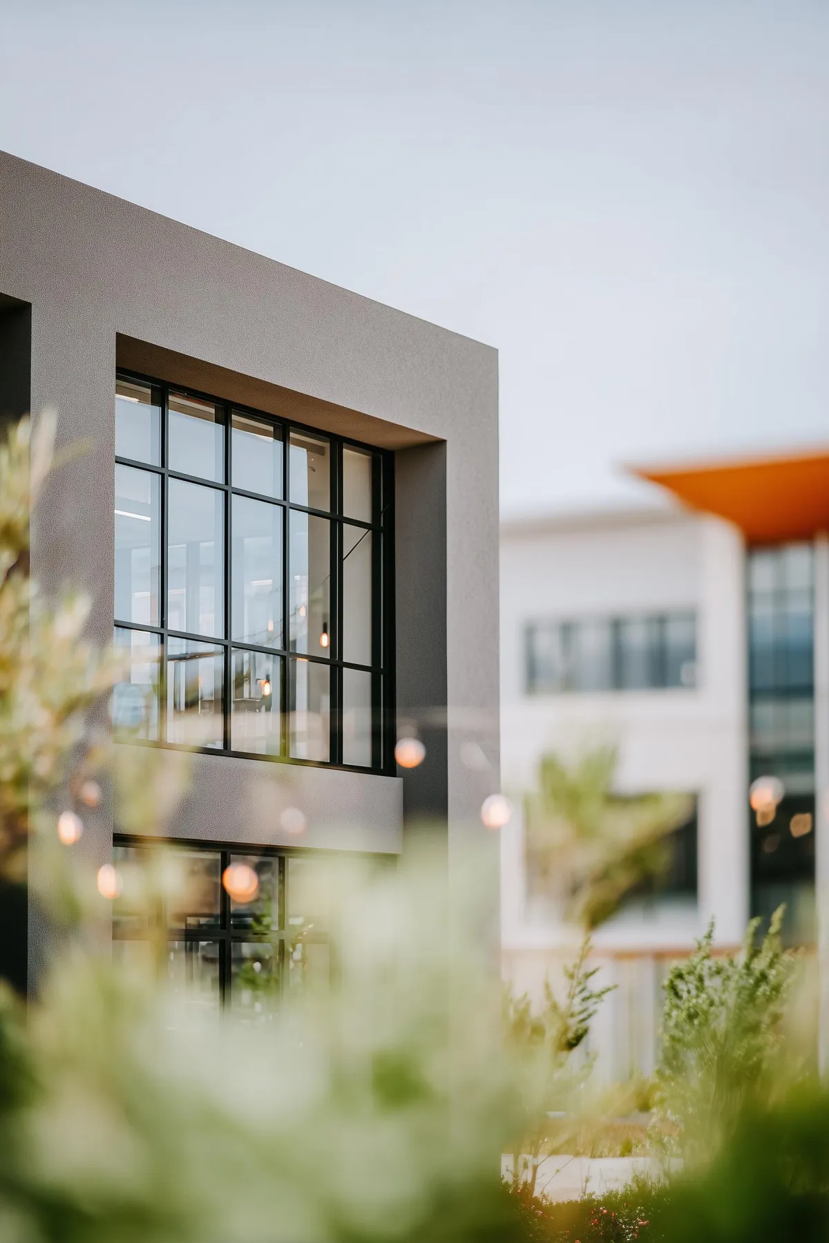 Modern commercial building exterior with large black-framed windows, clean gray architectural lines, soft bokeh lighting, and blurred greenery in the foreground, creating a contemporary and welcoming business campus setting.