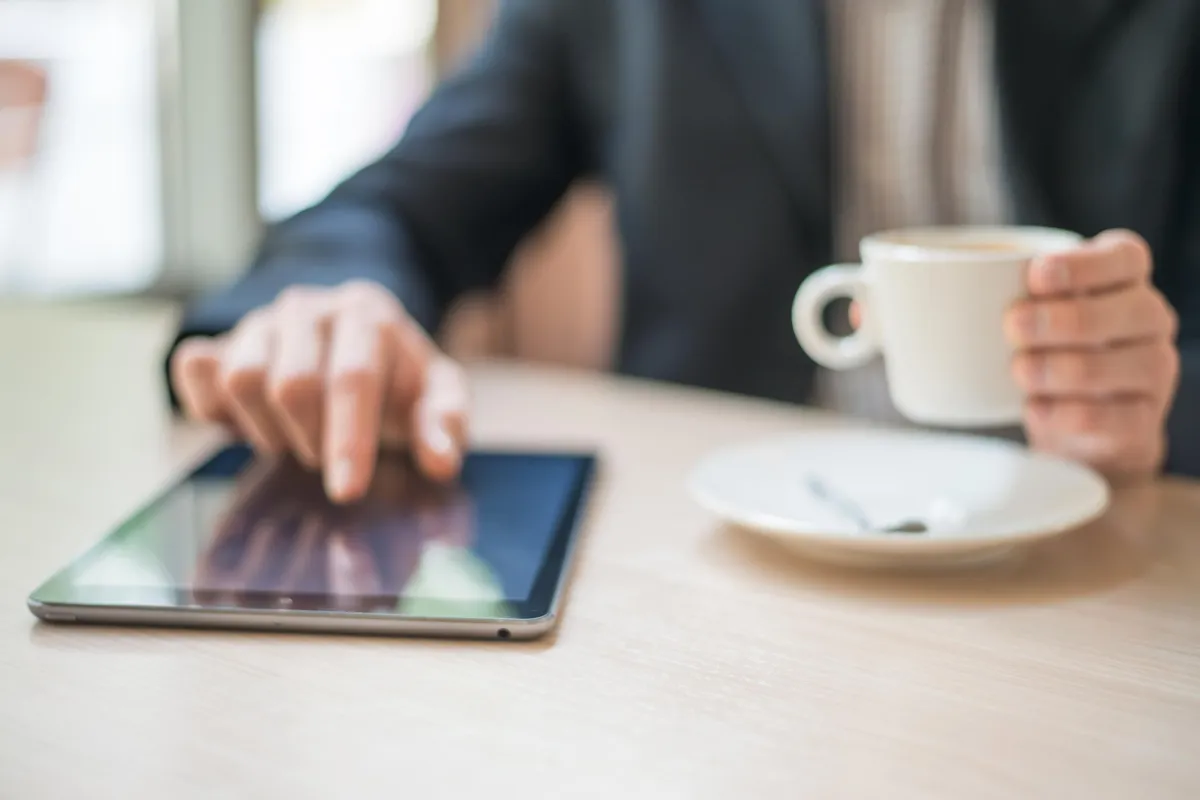 A person in a business suit using a tablet with one hand while holding a cup of coffee at a table, with a saucer and blurred background in a cafe setting.