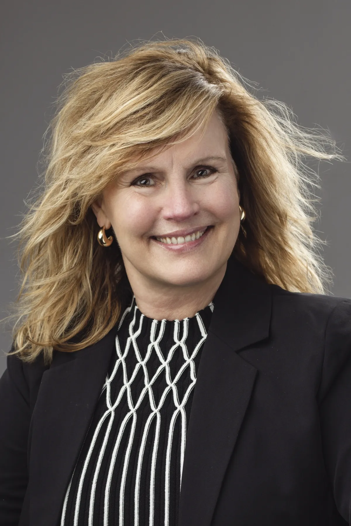 Smiling professional woman with curly blonde hair, wearing a black blazer and striped top, posing for a portrait against a neutral background.