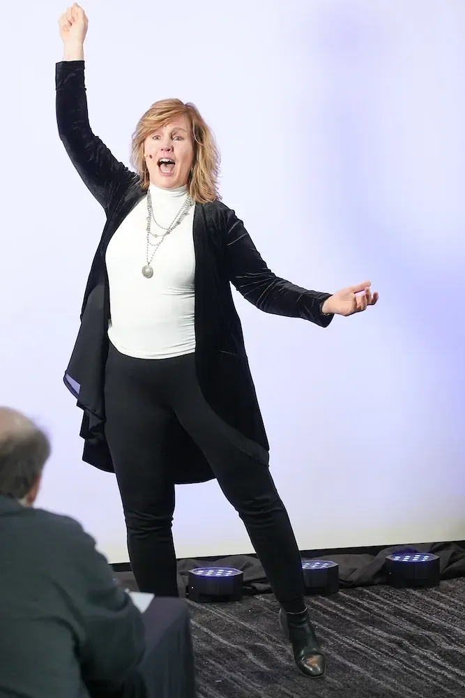 Energetic woman with blonde hair, wearing a black jacket and leggings, raising her arm in a victory pose during an event presentation.