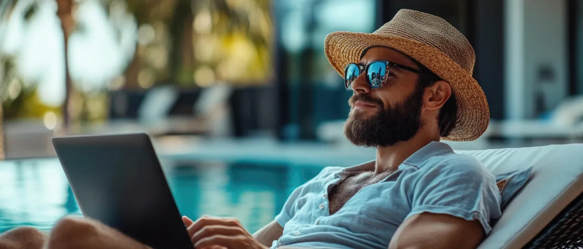 A man in his 40s, relaxed in casual clothes, closes his laptop at a kitchen table, looking out a window at a sunny backyard, symbolizing freedom from commuting and a peaceful work environment.