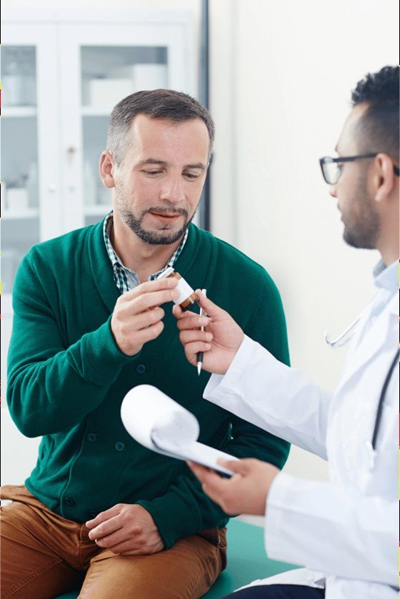 A male doctor in a white lab coat holding a small vial, consulting with a younger male patient in a clinical setting