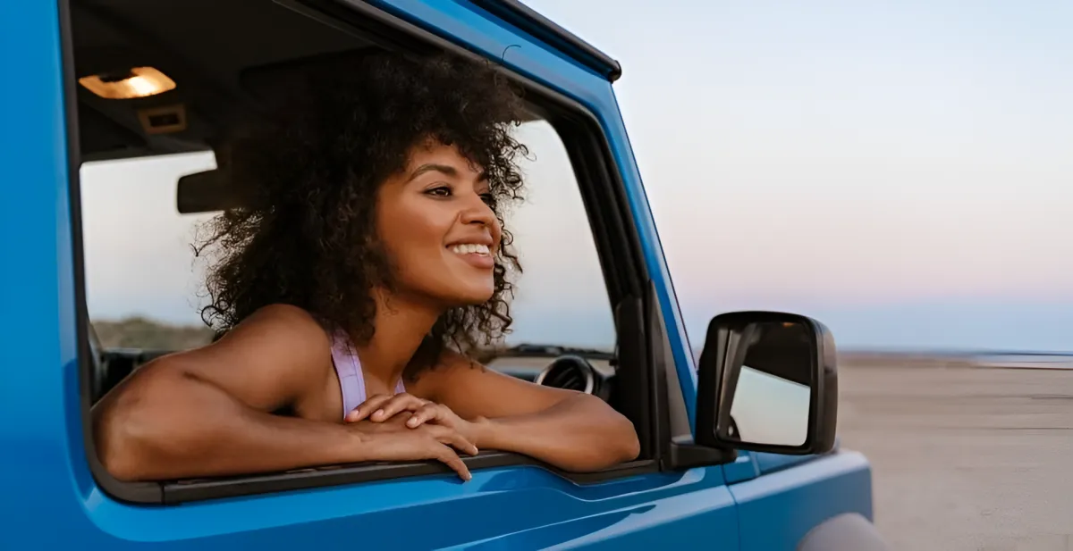 A woman smiles while sitting and looking outside from the driver's seat window of a blue car.