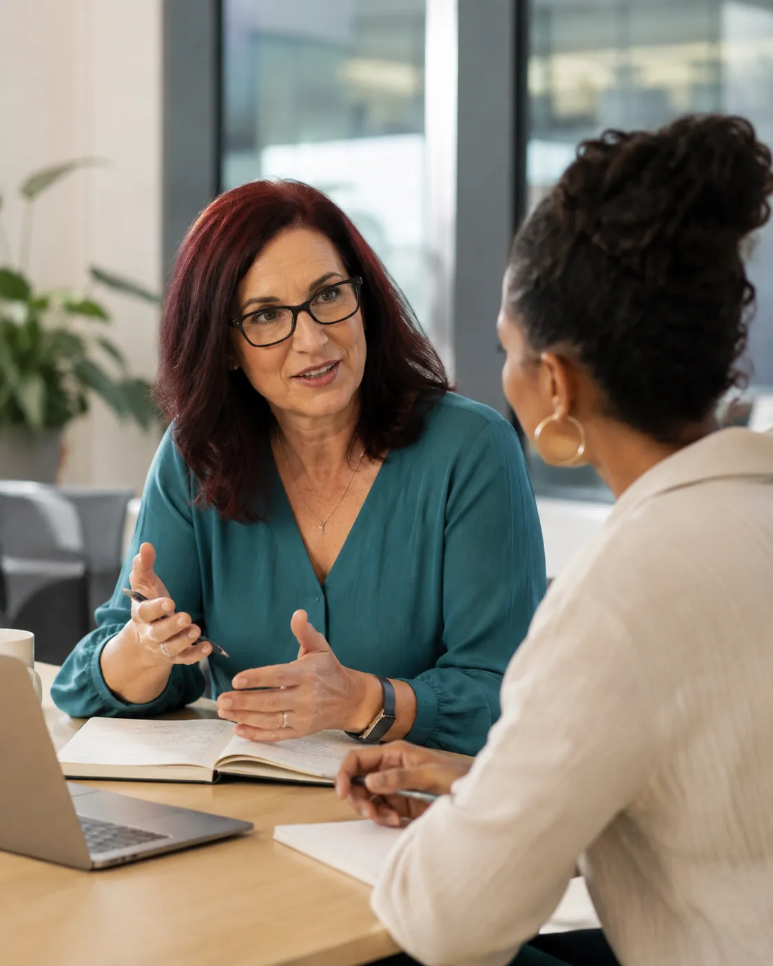 Woman in her late 50s with darker red hair, glasses, and a red coat in a candid leadership coaching conversation with a suited leader