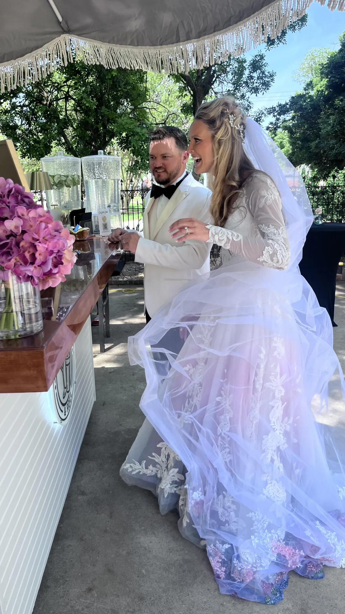 Bride and groom smiling at the Shake Rattle & Roll mobile bar during their outdoor wedding, with a professional bartender serving drinks under a fringe umbrella
