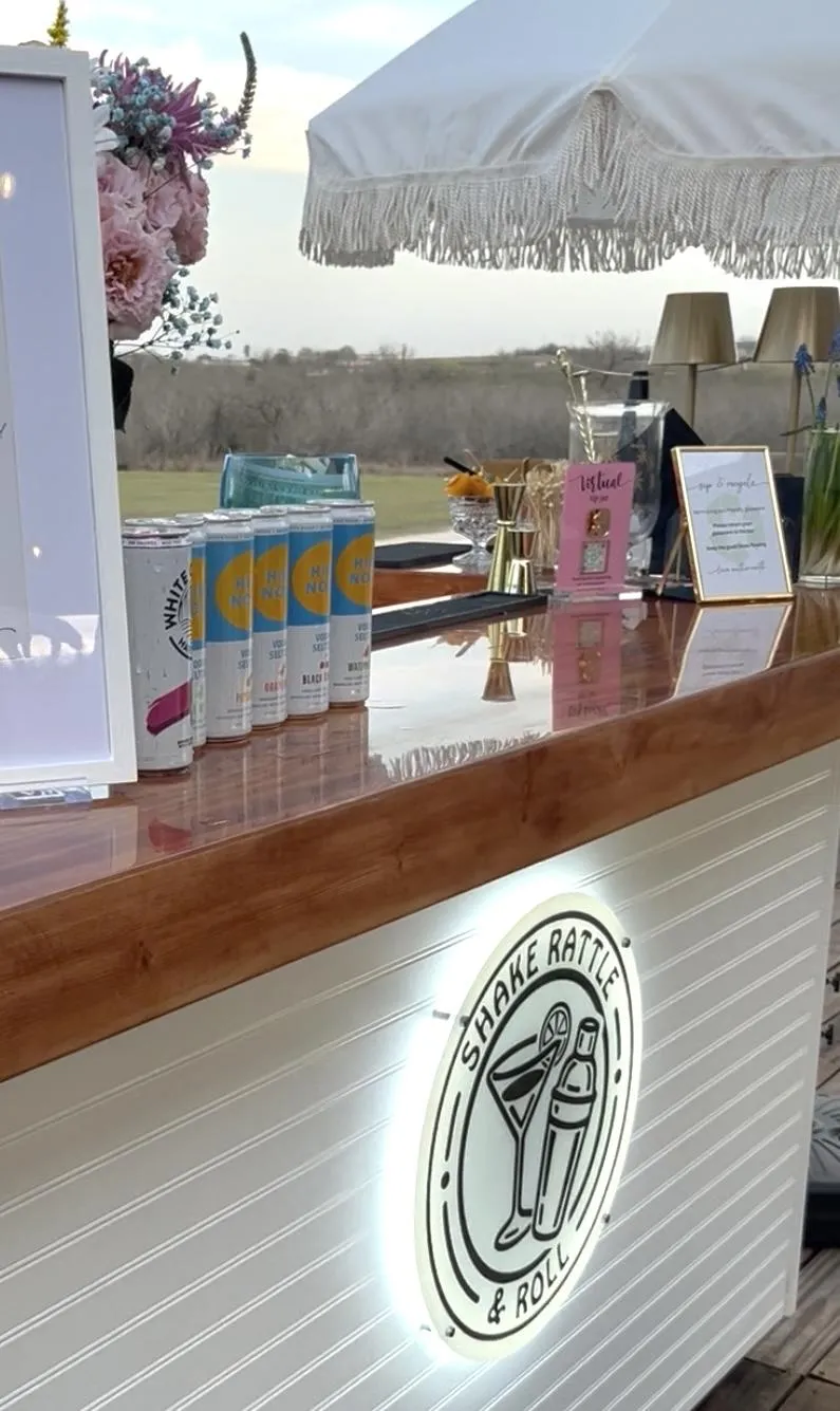 Front view of the Shake Rattle & Roll mobile bar featuring the Rollin Package with beer and wine service, canned beverages displayed on the bar under a fringe umbrella at an outdoor Texas event