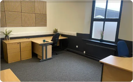 A furnished office with wooden desks, blue office chairs, gray carpet, black and cream walls, a large window, potted plants, and geometric wood wall art.