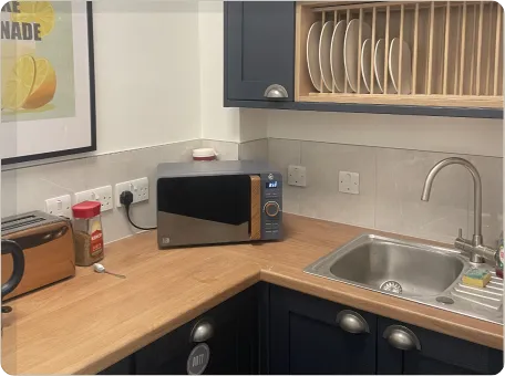 A small modern kitchen corner with navy cabinets, wood countertops, a stainless steel sink, a microwave, toaster, and wall-mounted plate rack. A framed lemonade print hangs on the left wall.