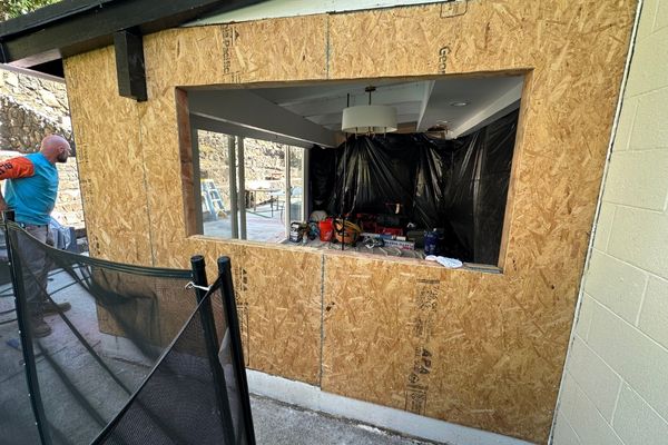A friendly local contractor consulting with a Hawaiian homeowner inside a bright living room, reviewing window and door options on a tablet. The room features tropical decor, natural wood finishes, and sunlight streaming through large, secure windows, conveying trust and expertise.
