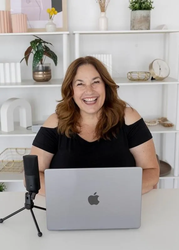 Jen Vazquez sitting at a white desk with white bookcases and her podcast mic and apple MacBook Pro in a black shirt smiling at the camera