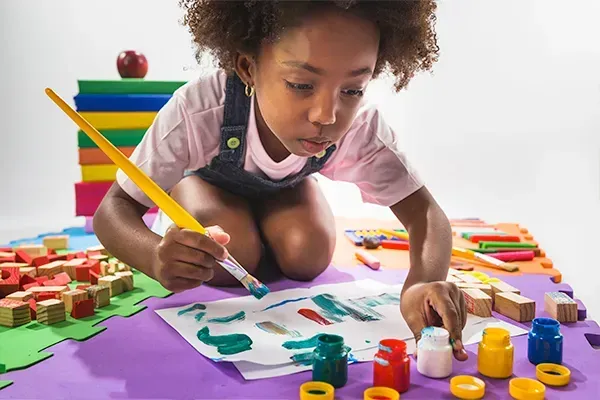 A young girl intently paints colorful strokes on paper using a brush and multiple paint jars around her.