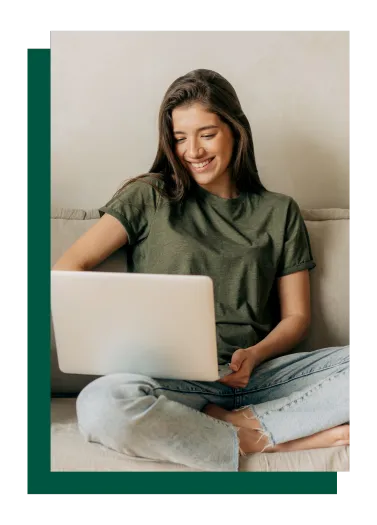 Woman with brown hair wearing a green shirt and jeans, smiling and laughing while looking at her laptop, seated on a cream couch with a distinctive green trim to one side of the photo