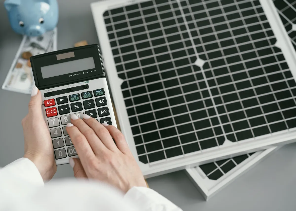 Person using a calculator next to small solar panels, dollar bills, and a piggy bank, symbolizing solar financing, savings, and return on investment.