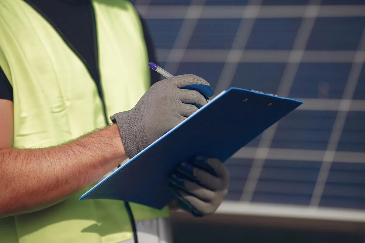 Close-up of a solar technician wearing safety gloves and a reflective vest, holding a clipboard and pen while performing a solar panel inspection.