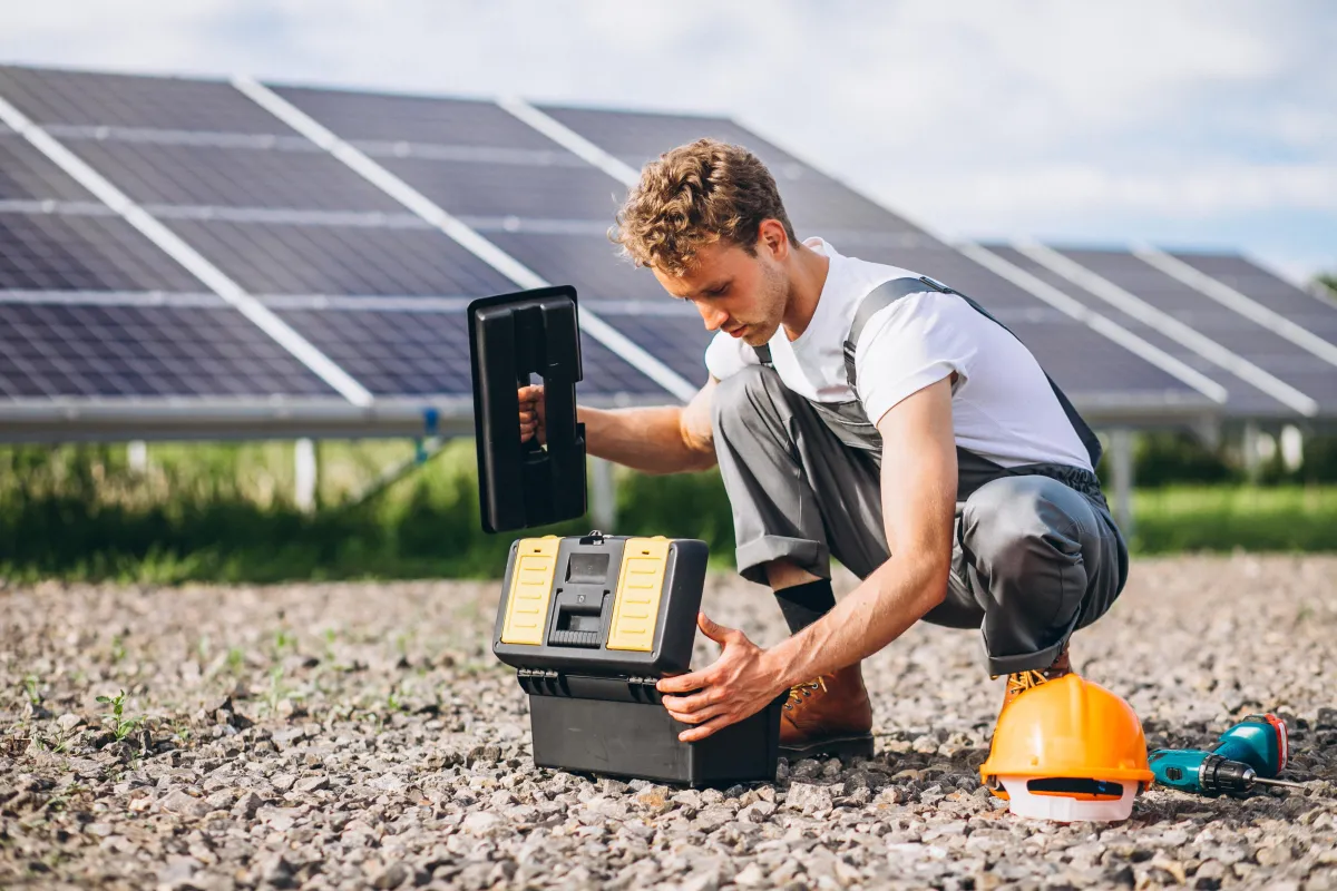 Technician kneeling beside an open tool box near a large ground-mounted solar panel array, with a hard hat and cordless drill resting nearby on the gravel.