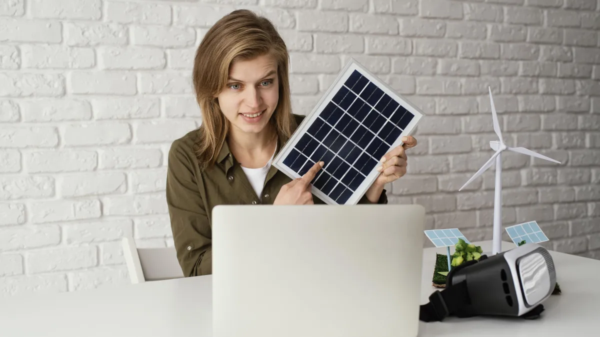 Woman sitting at a desk holding a small solar panel in front of a laptop, with miniature wind turbines and solar models nearby, demonstrating interest in renewable energy and sustainability.