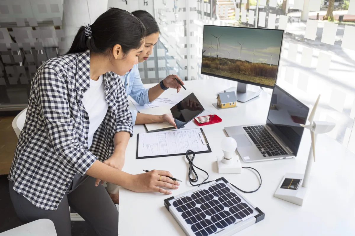 Two women working at a desk with solar and wind energy models, a laptop, and tablet, reviewing documents and digital data related to renewable energy planning or project design.