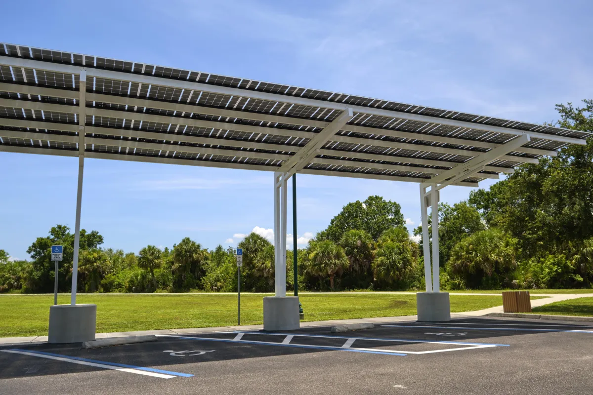 Solar carport structure installed over an open parking area with designated handicap spaces, featuring rooftop solar panels under a clear blue sky near a grassy park.