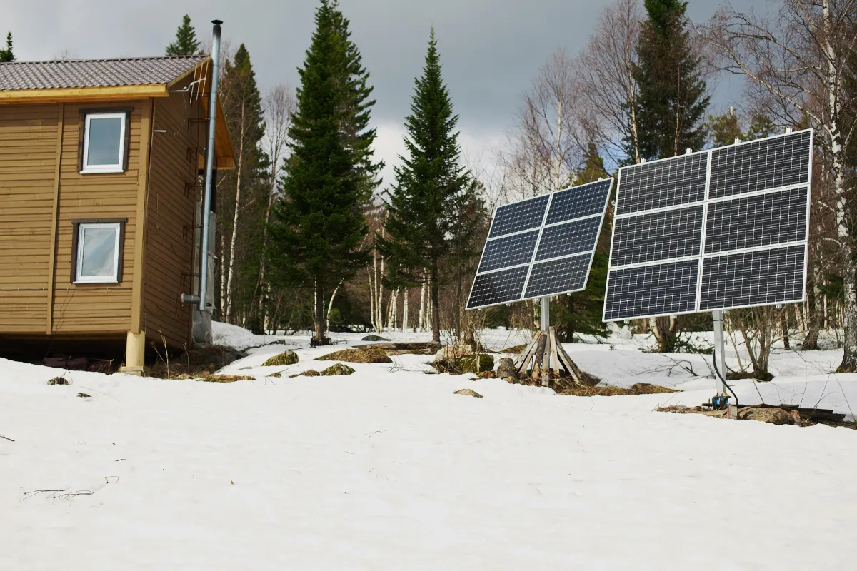 Off-grid cabin in a snowy forest with two ground-mounted solar panel arrays positioned nearby, surrounded by pine trees under a cloudy sky.