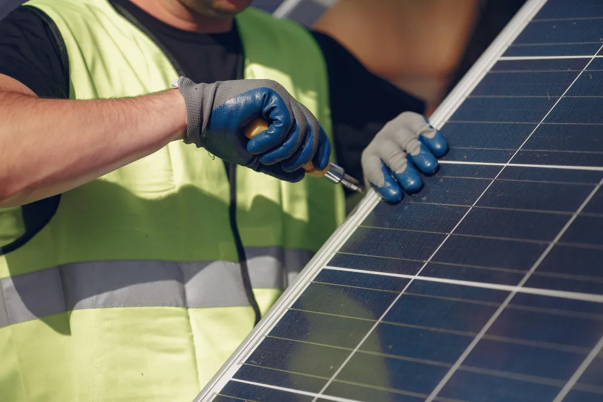 Close-up of a solar technician in a high-visibility vest and gloves securing a solar panel with a hand tool during installation or maintenance.