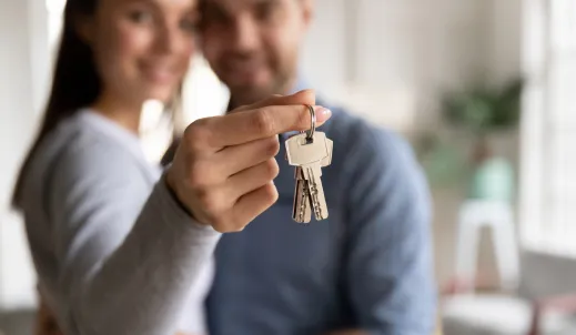 "A happy couple standing in a bright, modern home, holding up a set of house keys towards the camera. The woman's hand is in focus, showcasing the keys, while the couple is slightly blurred in the background, smiling. The image conveys themes of homeownership, real estate, and new beginnings.