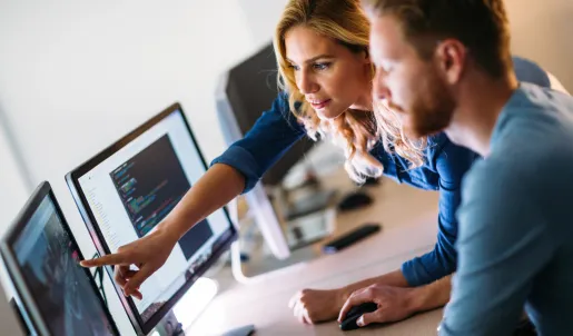 Two professionals, a woman and a man, collaborating at a workstation with multiple computer monitors. The woman, wearing a denim shirt, is pointing at a screen displaying lines of code, while the man, dressed in a dark blue sweater, attentively follows along. The office setting is modern, with dim lighting and a focus on the illuminated screens, suggesting a tech, software development, or data analysis environment.