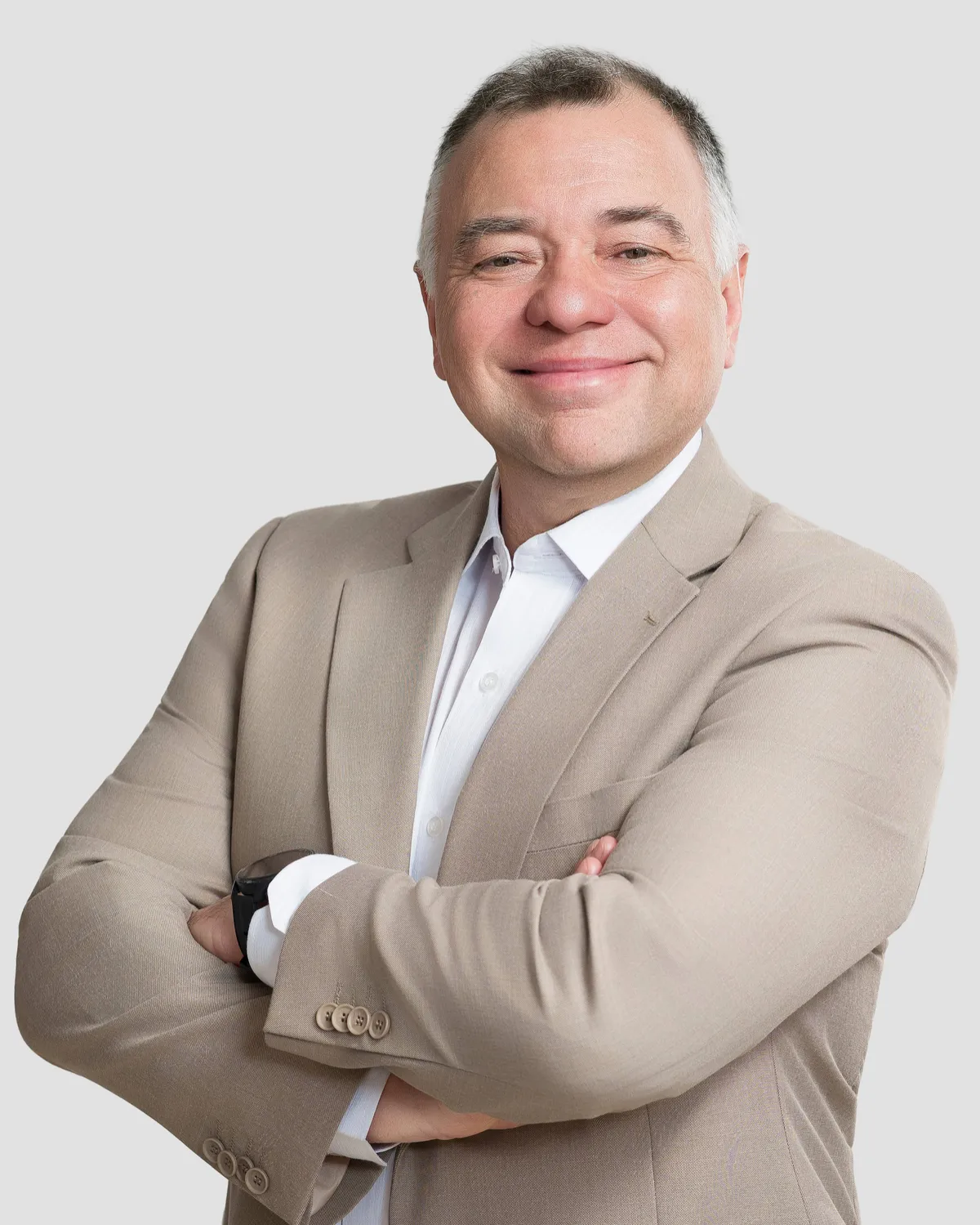 Professional portrait of the same man, now wearing a tan blazer and a light shirt, standing with arms crossed