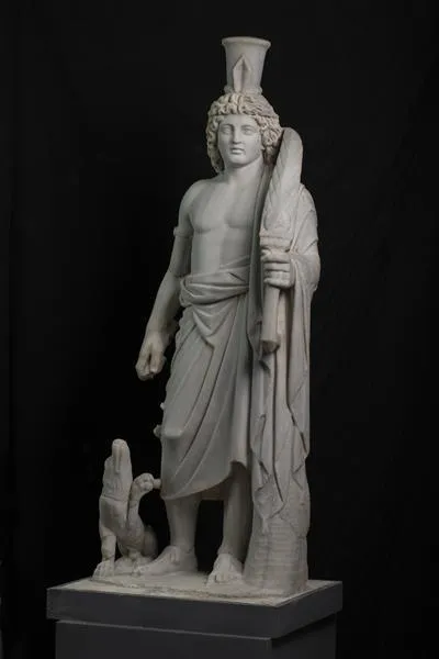 Hermanubis  This statue represents a draped athletic male figure with his sandaled feet standing on an integral plinth and resting against a tree trunk which serves as a support. He is identified as a new iconographic type, introduced into the pantheon of Roman Alexandria, and generally called Hermanubis, despite the caveat raised by Benaissa.  This god is a syncretistic deity in whom the characteristics of Anubis, the jackal-headed god of embalming, and those of Hermes, the Greek god charged with leading souls to the hereafter, are combined. He is clean-shaven with a full head of thick curly locks on which he wears a modius fronted by a stylized palm frond. His left hand holds a second, more clearly defined, palm frond set into a sword-like hilt emblazoned with a circular device. The attribute in his lowered right hand may very well have been a caduceus, the top of which has attracted the attention of his accompanying dog, clearly intended as an allusion to the jackal with whom Hermanubis was associated. The substitution of a dog for a jackal may have been motivated by the association of Anubis with  the dog in numerous Greek texts.     From Ras el-Soda Temple, now at the Antiquities Museum of Bibliotheca Alexandrina