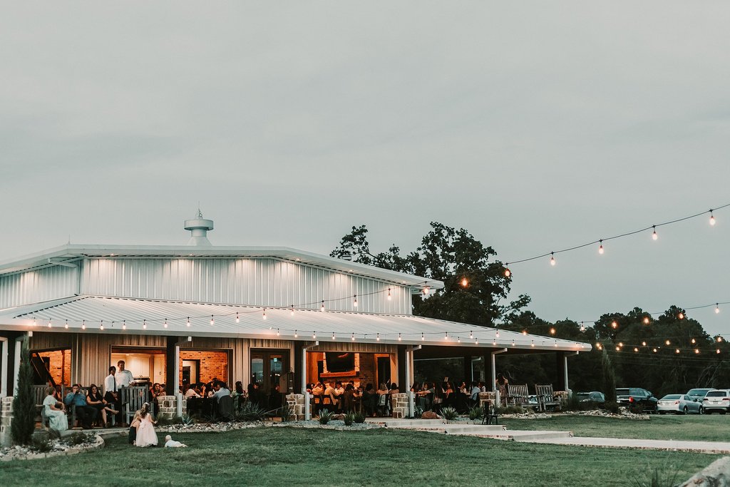 Aerial view of Twisted Pines Winery estate with rolling vineyards, a rustic-modern tasting room, and a serene pond reflecting the sky, all set against distant hills and a soft, golden sunrise.