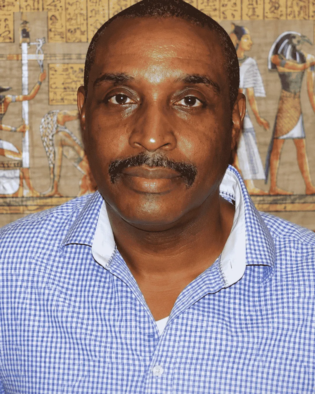Portrait of Lalji Selassie, founder of Ascension Knowledge, wearing a blue checkered shirt with an ancient Egyptian-themed background representing spiritual wisdom and conscious transformation.