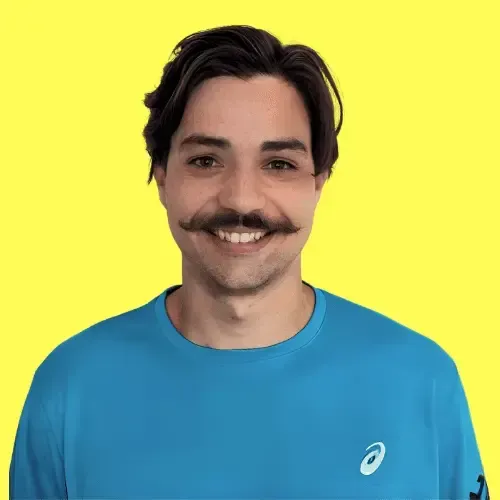  A smiling man with dark wavy hair and a thick mustache, wearing a bright blue Asics sports shirt, standing in front of a solid yellow background.