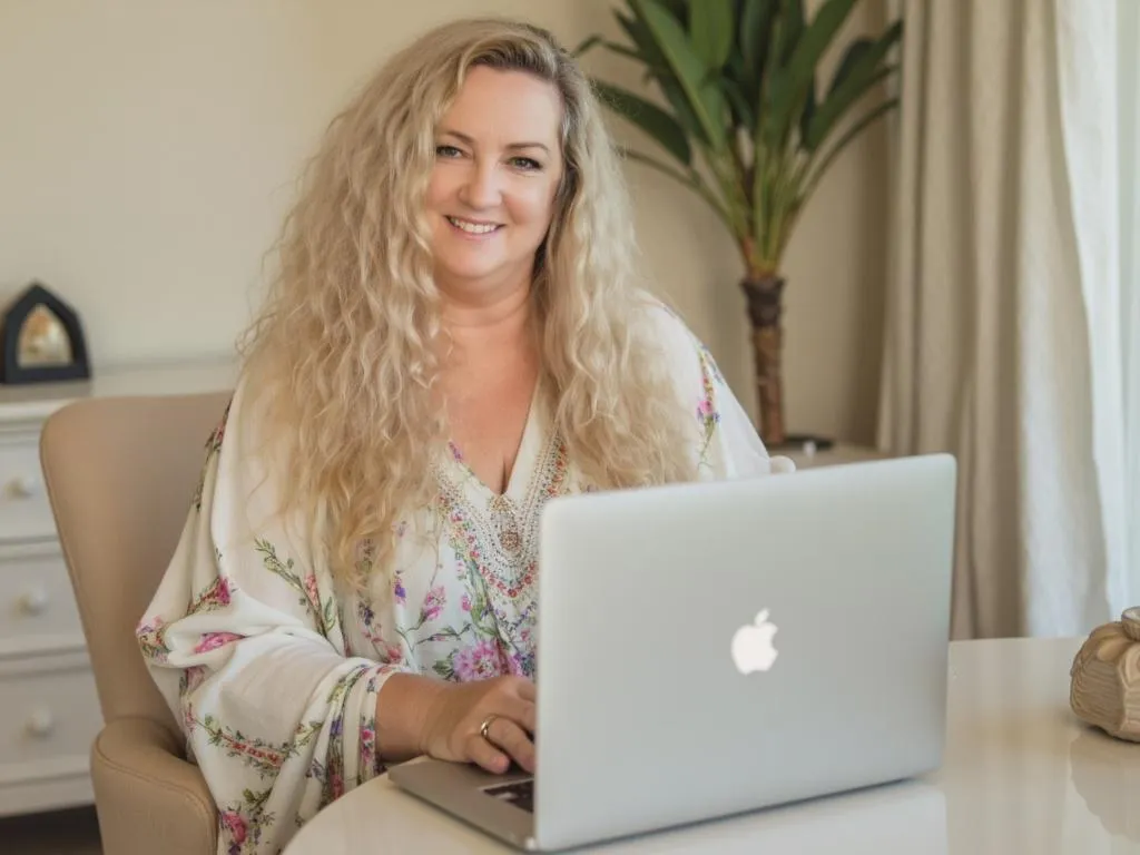 This image shows a Debbie Mewes CEO of Gem Software & Coaching with long, curly blonde hair sitting outdoors, focused on a laptop in front of her. She is wearing a flowing blue and white patterned top. The background features a lush, sunny setting with green trees, vibrant orange flowers, and a serene pool or water feature, suggesting a peaceful and natural environment. The overall mood is professional yet relaxed, blending work and leisure in a tranquil outdoor space