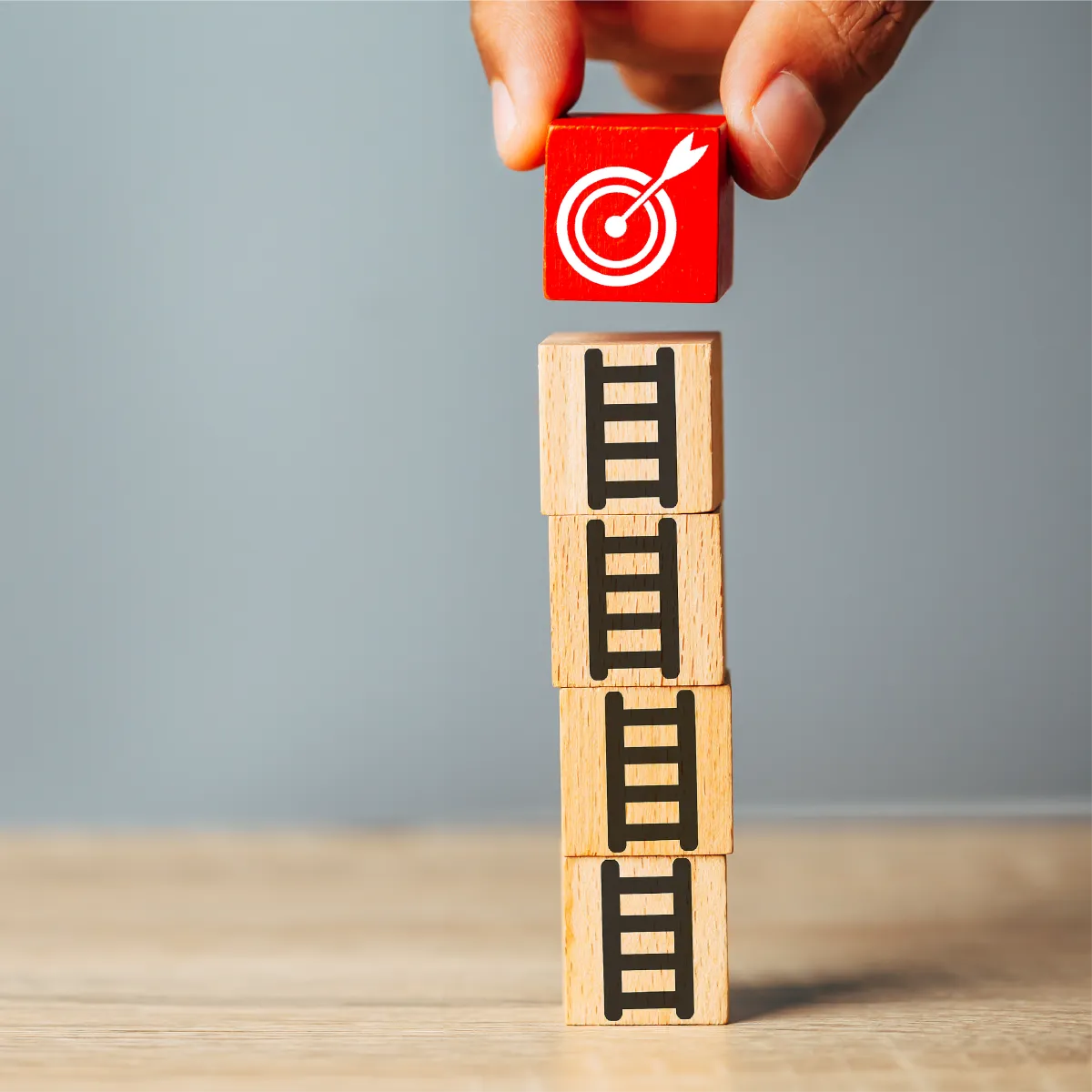 A hand places a red block with a target and arrow icon on top of a stack of wooden blocks with ladder icons, symbolizing the steps to achieving goals.