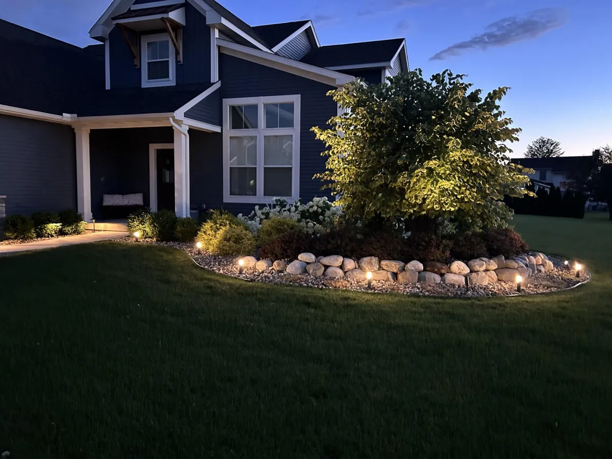 Front yard at dusk with pathway lighting, rock-bordered garden bed, shrubs and tree illuminated in front of a dark blue-gray modern house.