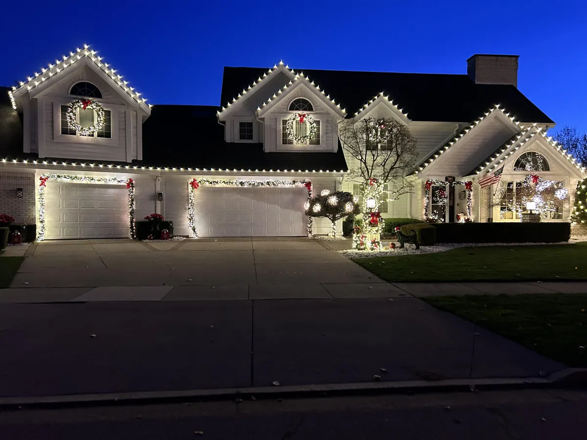 House at dusk decorated with white holiday lights outlining the roofline and garage, plus wreaths and lit garlands.