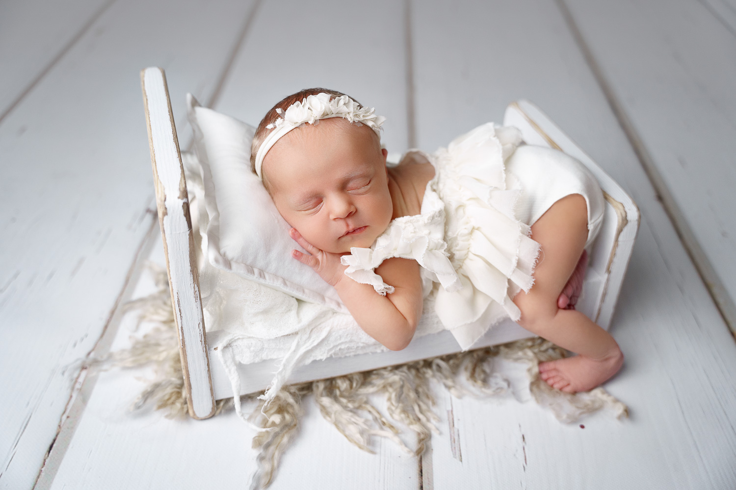 Babymädchen beim Babyshooting Hamburg posiert in Bauchlage auf eine weißen Bettchen aus Echtholz in weißem Babykleid und Stirnband stehend auf einem weißen Holzhintergrund