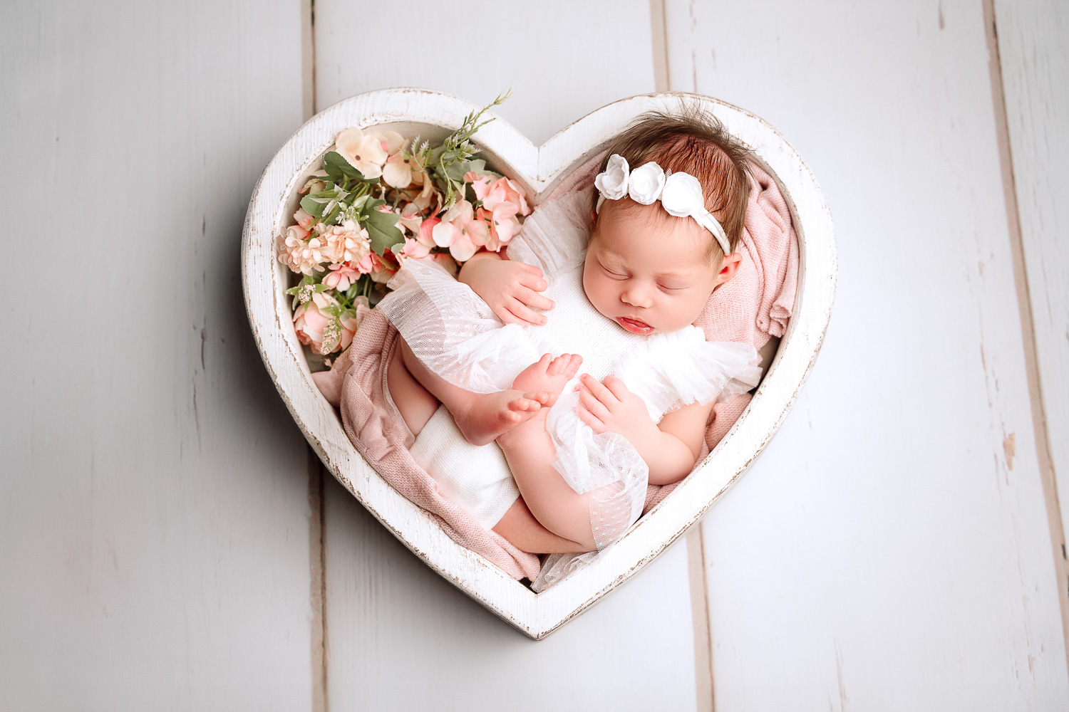 Baby in einer weißen Herzschale positioniert in einem weißen Ballerinakleid und passendem Stirnband.