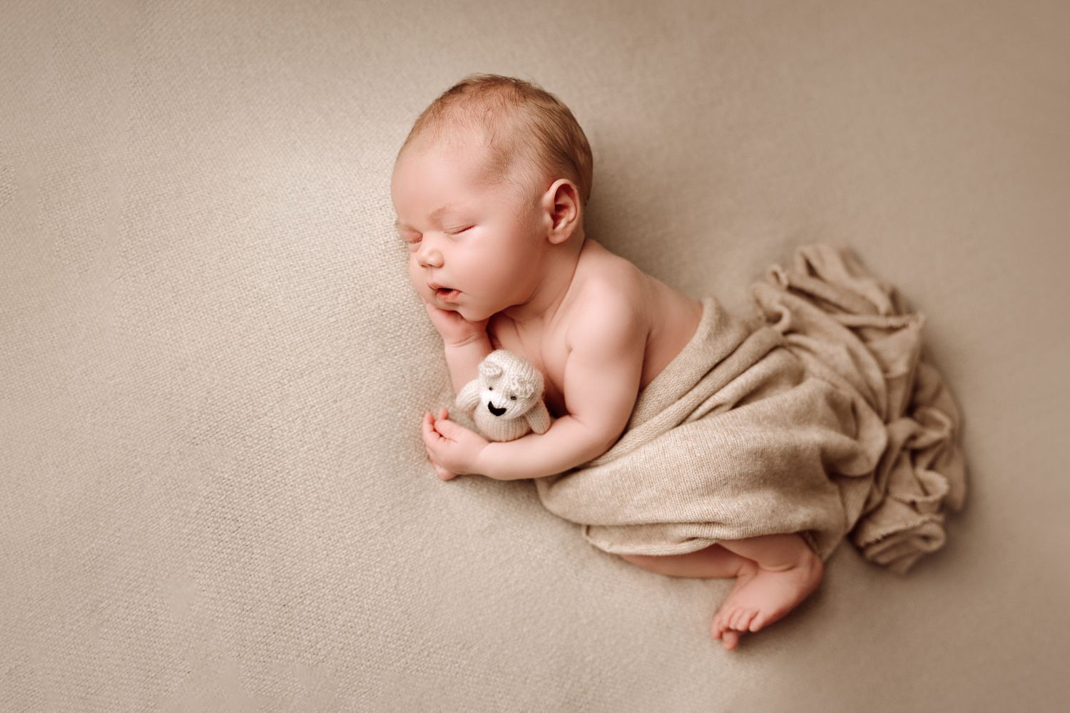 Baby auf einer Decke in der Farbe beige auf der Seite liegend mit einem Tuch bedeckt und einem Teddy im Arm beim Shooting Hamburg