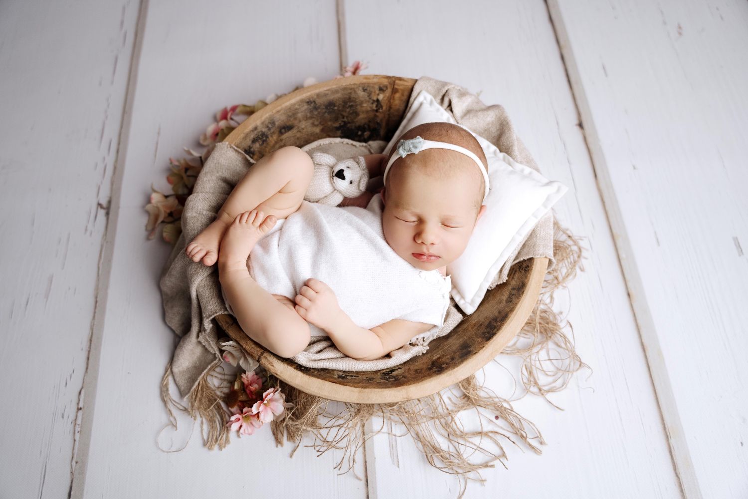 Baby Mädchen auf Babyfoto Hamburg auf dem Rücken liegend in einer braunen Echtholzschale, weiß gekleidet mit einem süßen Stirnband und Teddybär im Arm auf einem weißen Holzhintergrund